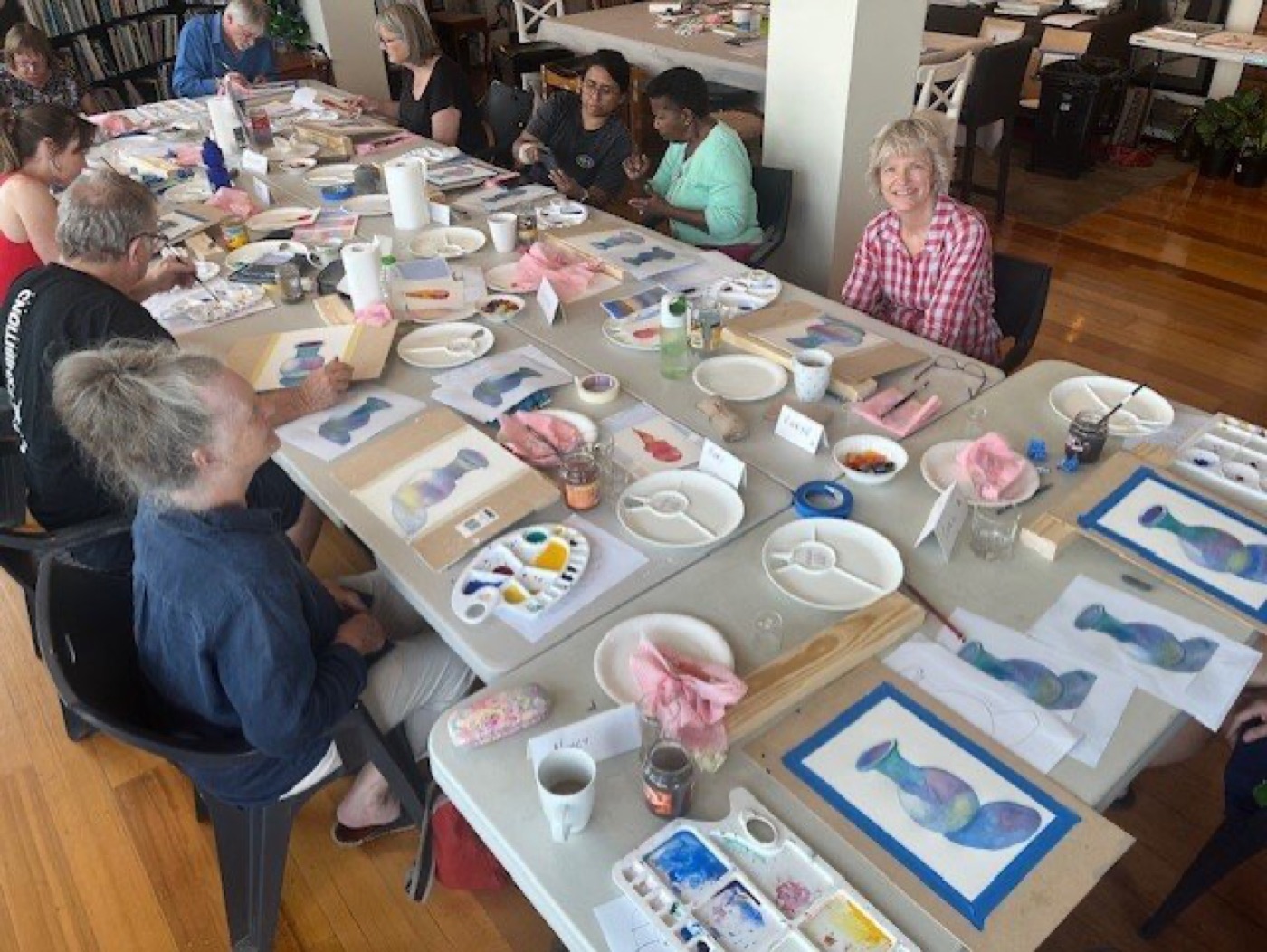 Students seated at a long table working on watercolour paintings of a vase